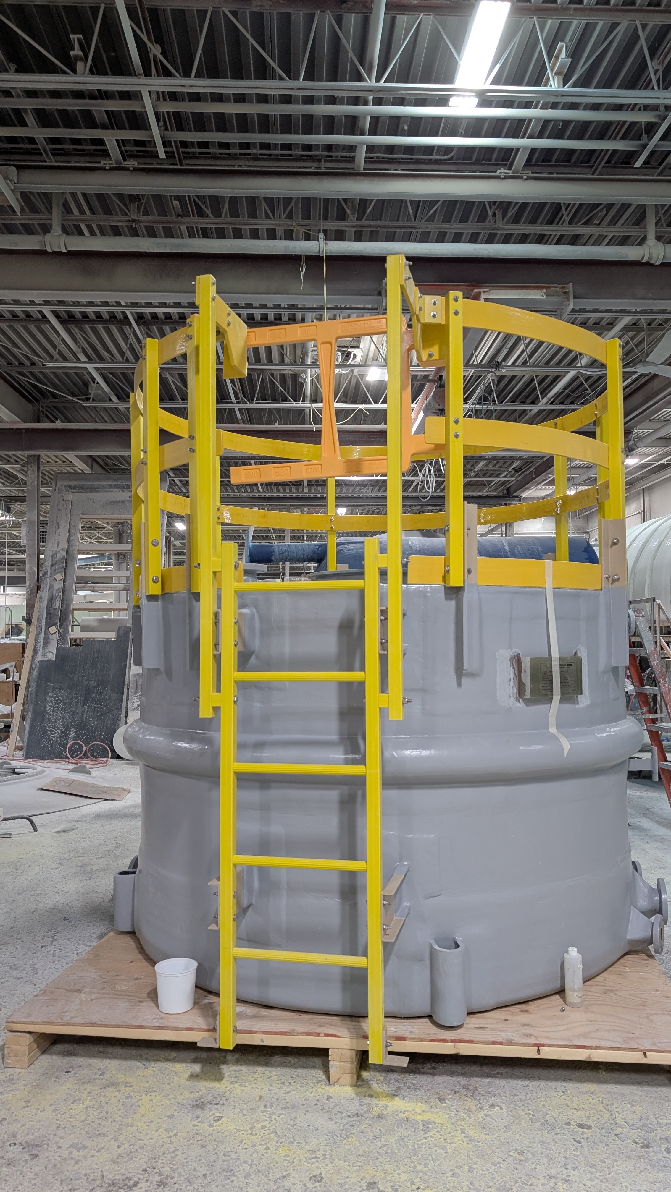 Yellow Intrepid UDG self-closing safety gate installed at the top of a vertical ladder on a large grey FRP tank in the Precisioneering shop